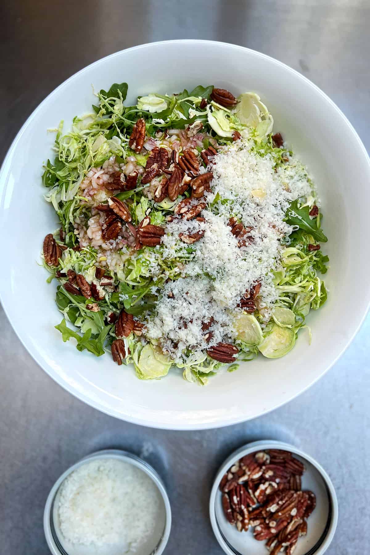 overhead shot of a white bowl filled with shaved brussels sprouts salad ingredients that haven't been tossed together yet: brussels, arugula, pecans, vinaigrette and parmesan