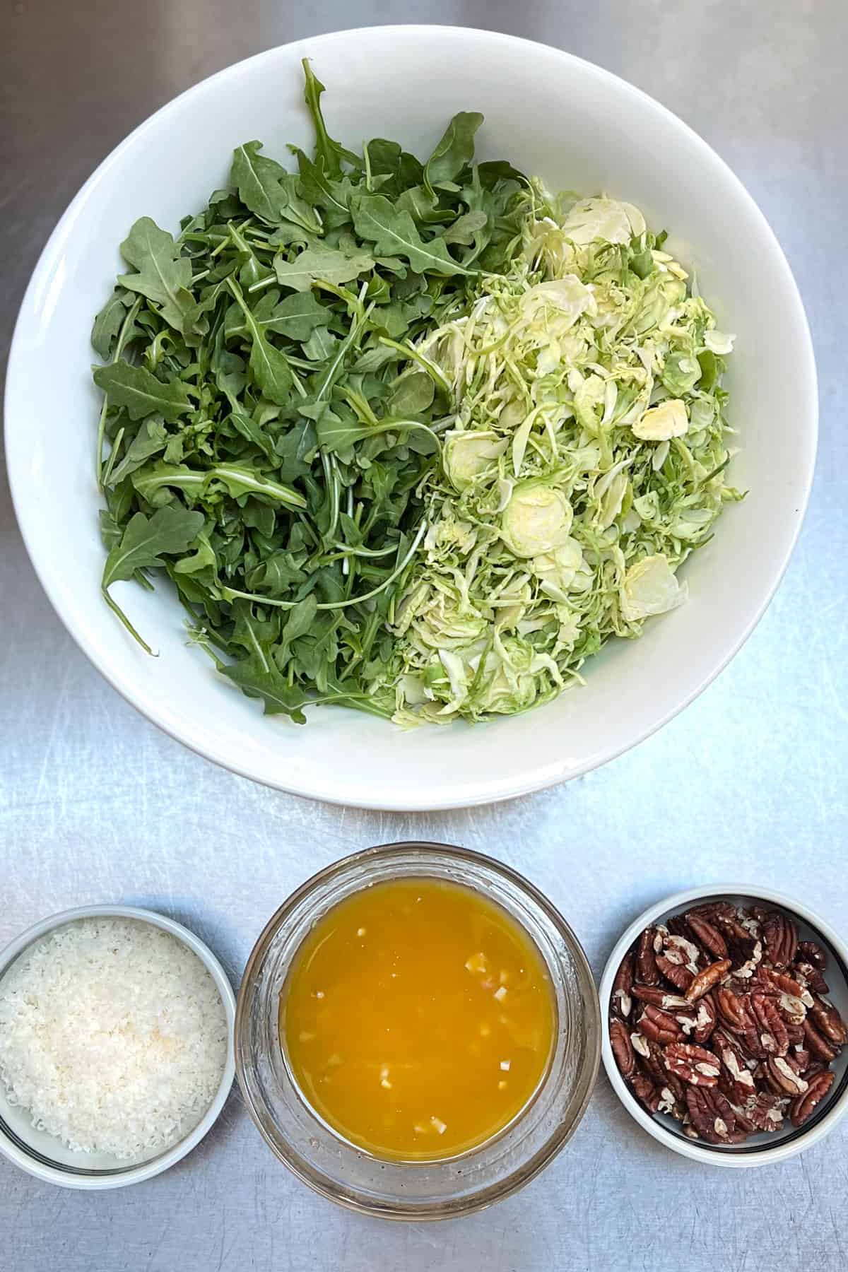 Overhead shot of a white bowl half filled with arugula the other half shredded brussels sprouts, three smalls bowls next to it with grated parmesan, vinaigrette, and toasted pecans