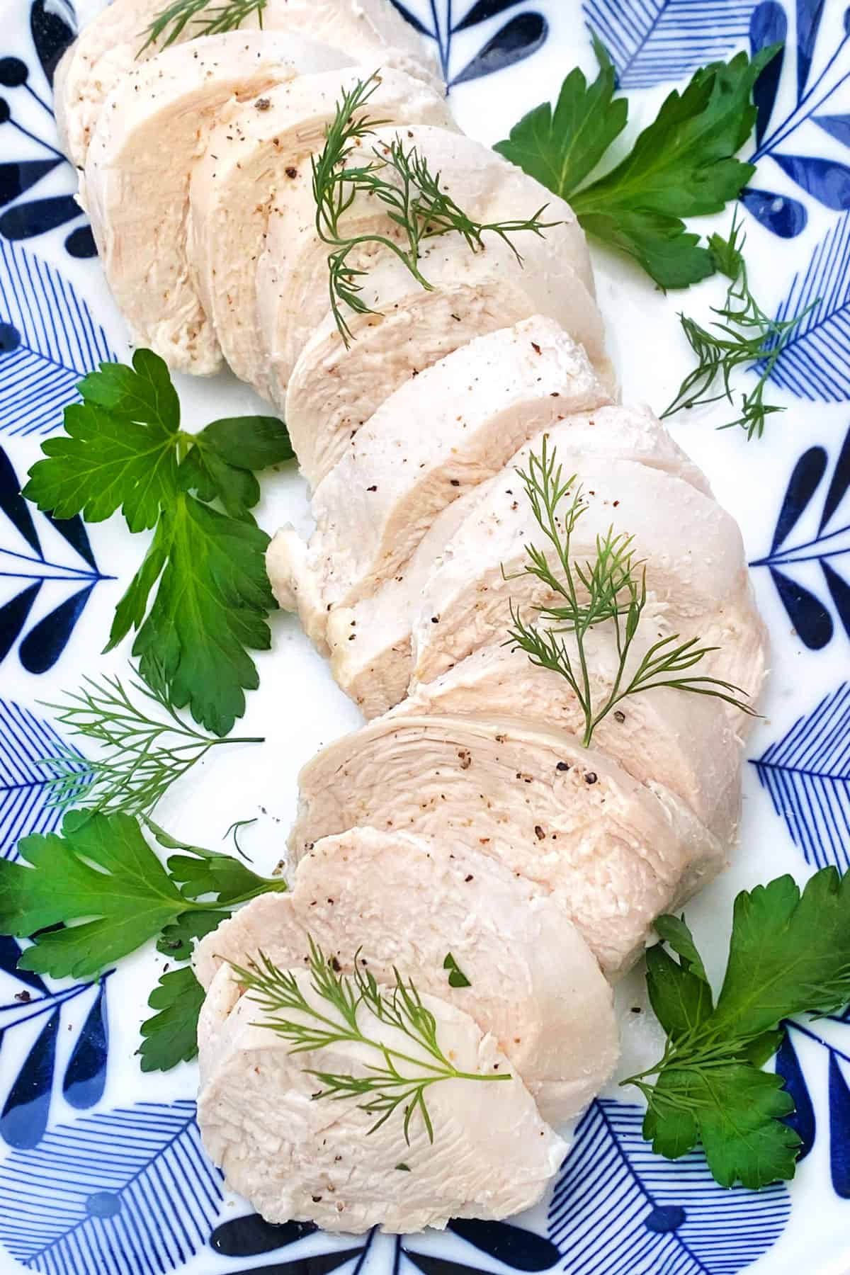 Close up of 5 slices of poached chicken on a blue and white plate, topped with some delicate sprigs of dill and parsley leaves