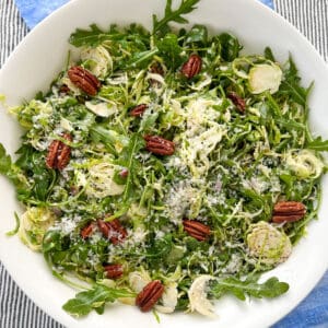 Overhead shot of a white bowl filled with shredded Brussels sprout and arugula salad with toasted pecans and grated parmesan cheese.