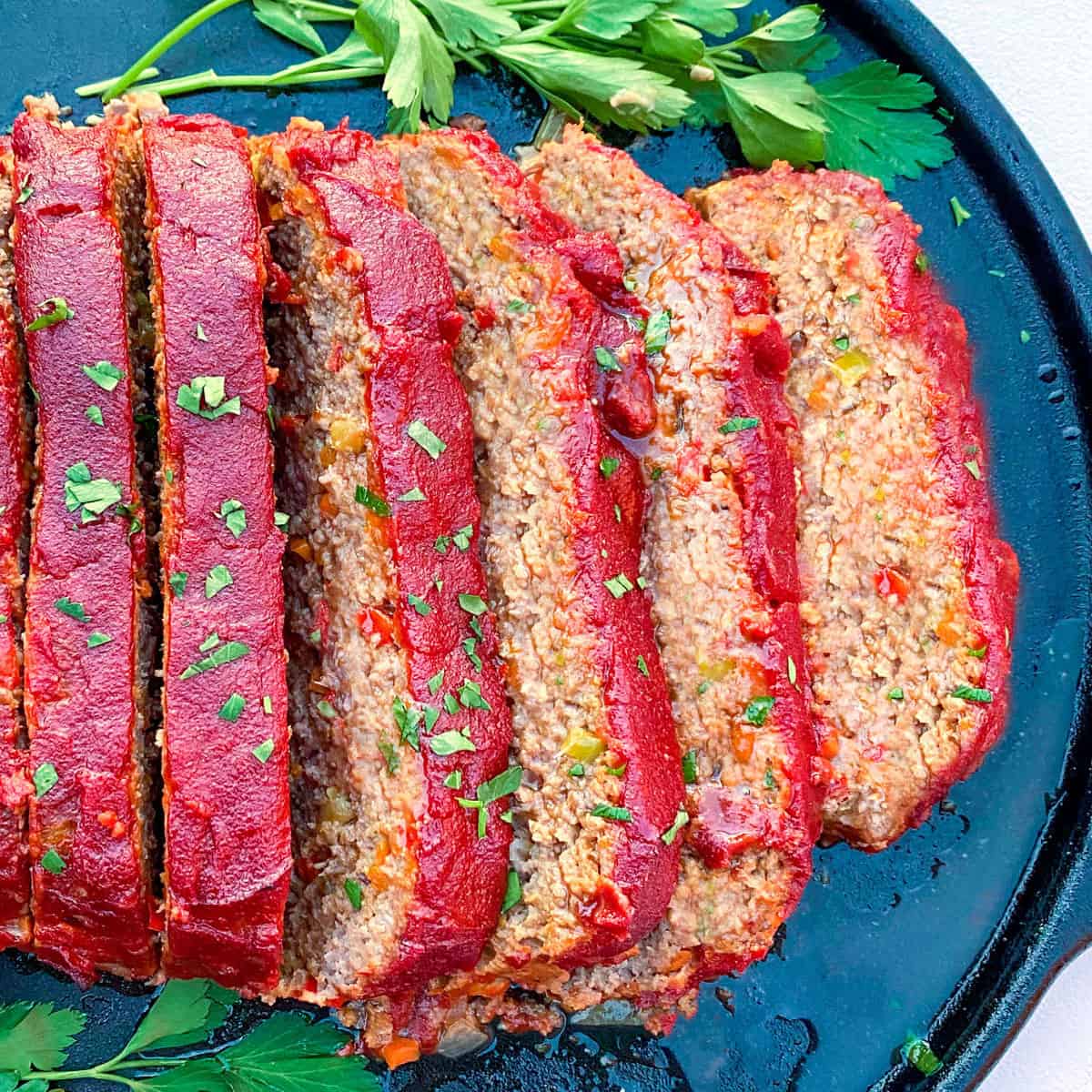 overhead shot of a tomato glazed meatloaf, sliced and set on a black platter and sprinkled with chopped parsley