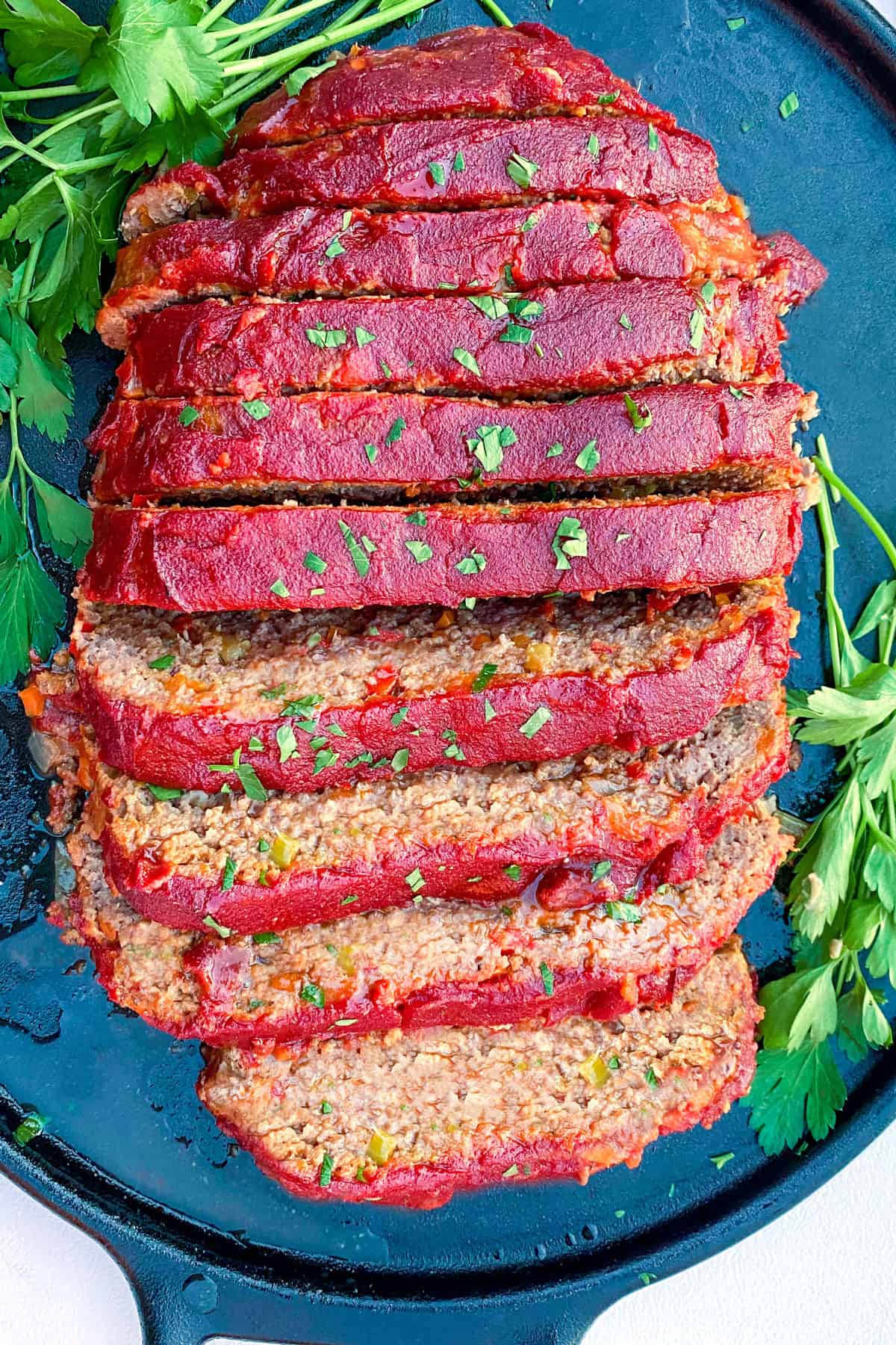 overhead shot of a tomato glazed meatloaf, sliced and set on a black platter and sprinkled with chopped parsley