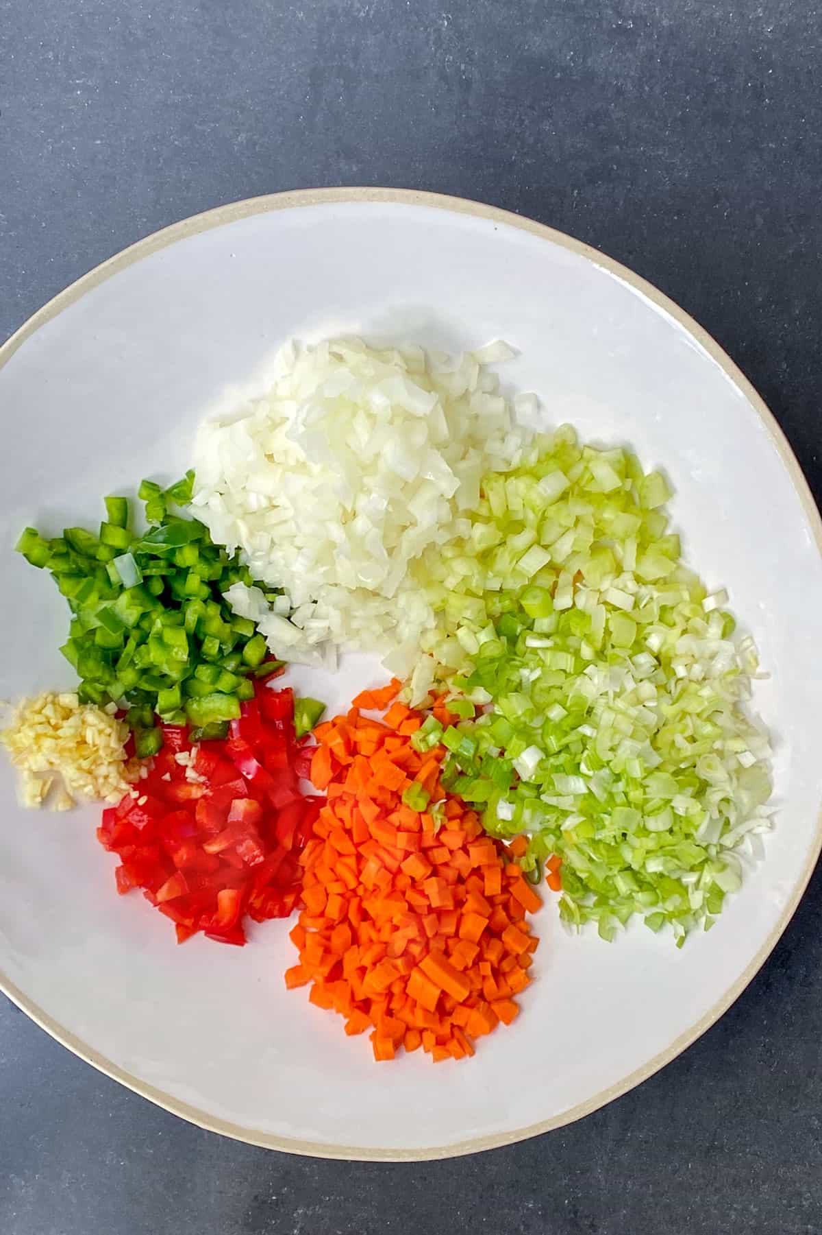 overhead shot of a large white bowl filled with piles of chopped veggies: onions, celery, carrots, red pepper, green pepper and garlic