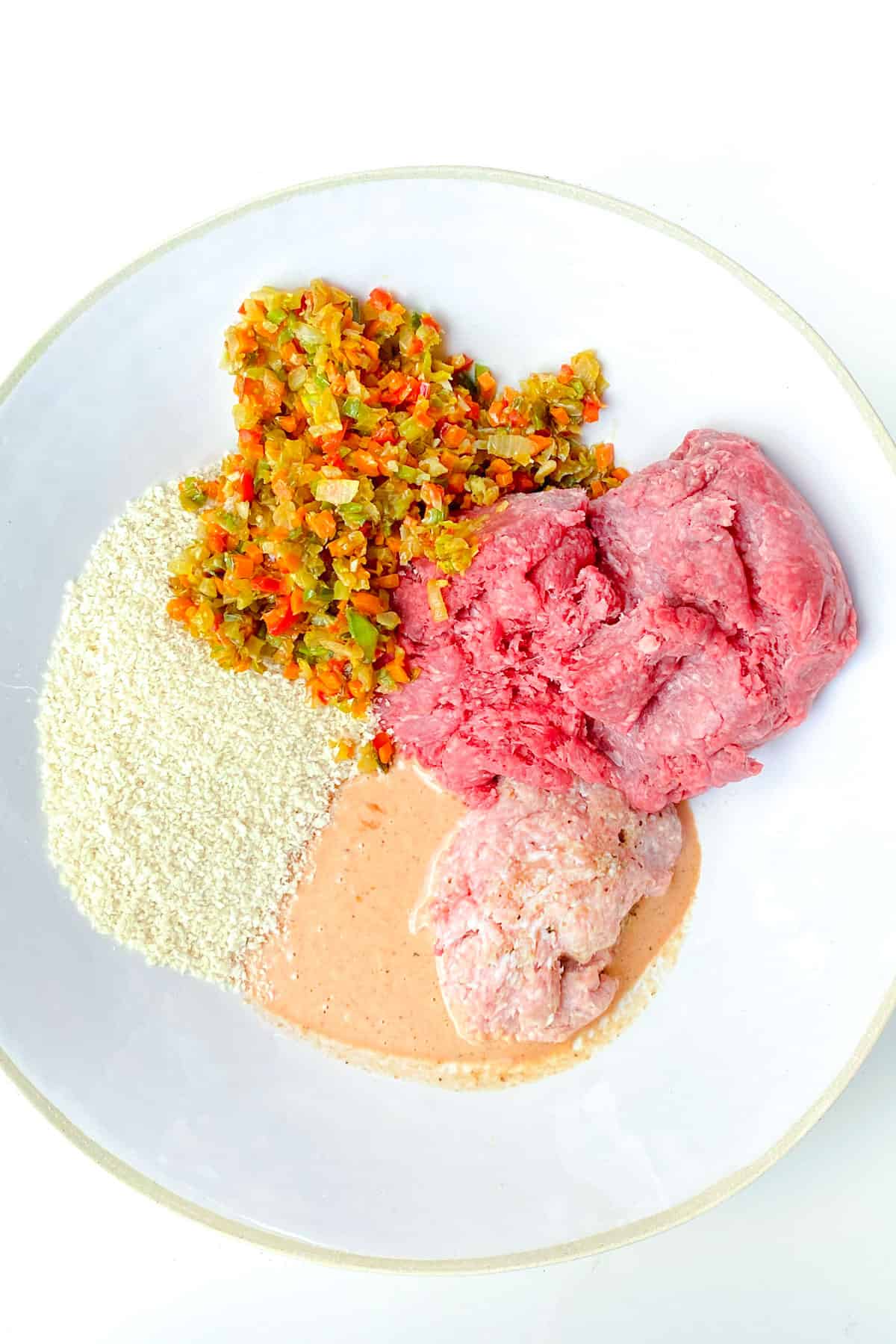 overhead shot of a white bowl filled with meatloaf ingredients: two kinds of ground meat, sauteed veggies, bread crumbs and a ketchup and cream mixture