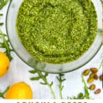 overhead shot of a glass bowl filled with arugula pesto, on a white countertop, surrounded by arugula leaves, 3 small piles of toasted pistachios and 3 lemons