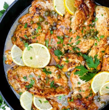 overhead shot of a black cast iron skillet filled with chicken piccata, thin chicken cutlets with a few thin slices of lemon tossed on top and some chopped parsley