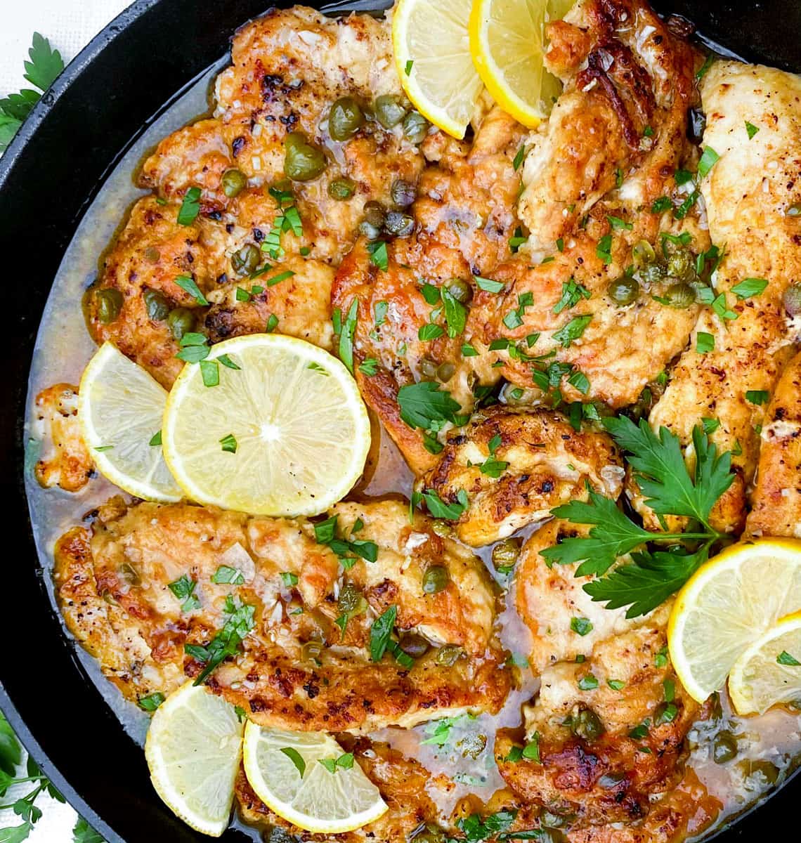 overhead shot of a black cast iron skillet filled with chicken piccata, thin chicken cutlets with a few thin slices of lemon tossed on top and some chopped parsley