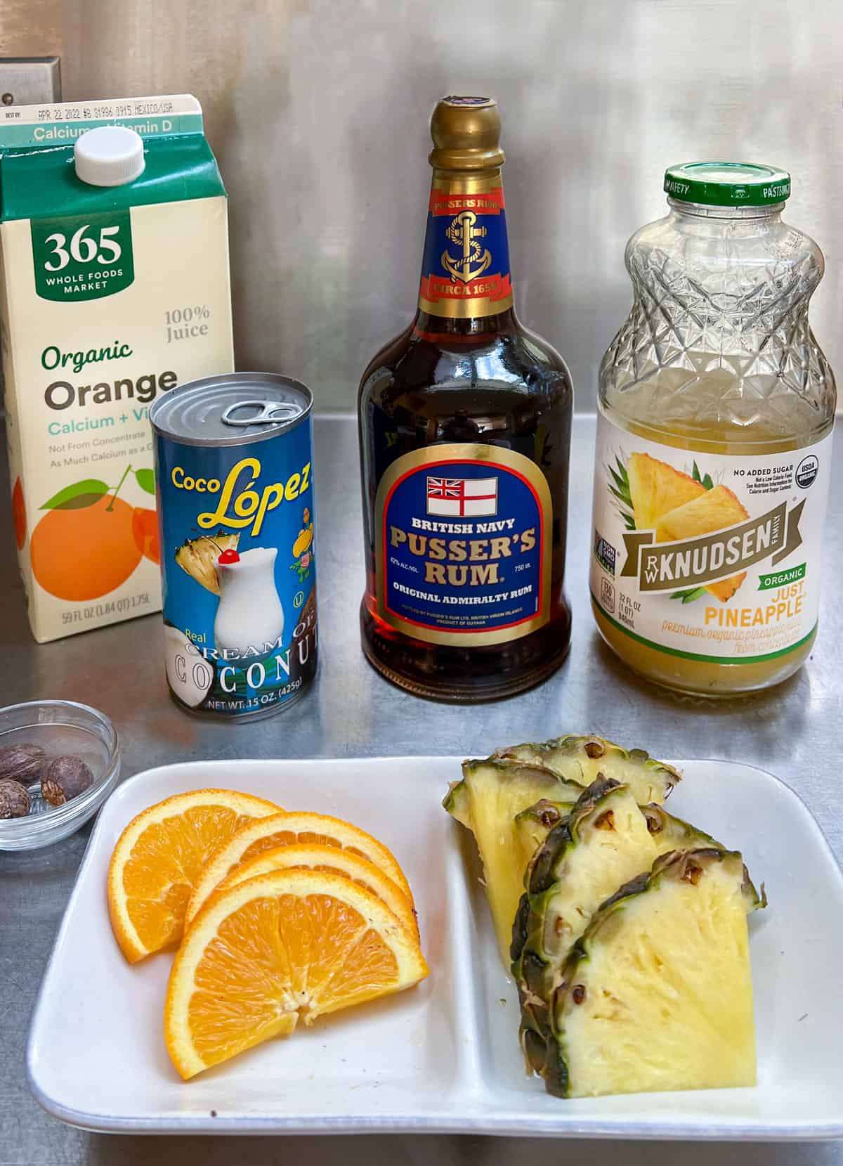 Kitchen counter with a container of orange juice, a bottle of Pusser's rum, a bottle of pineapple juice, a can of coco lopez and, in the foreground a small patter with thin orange slices and triangular pineapple slices