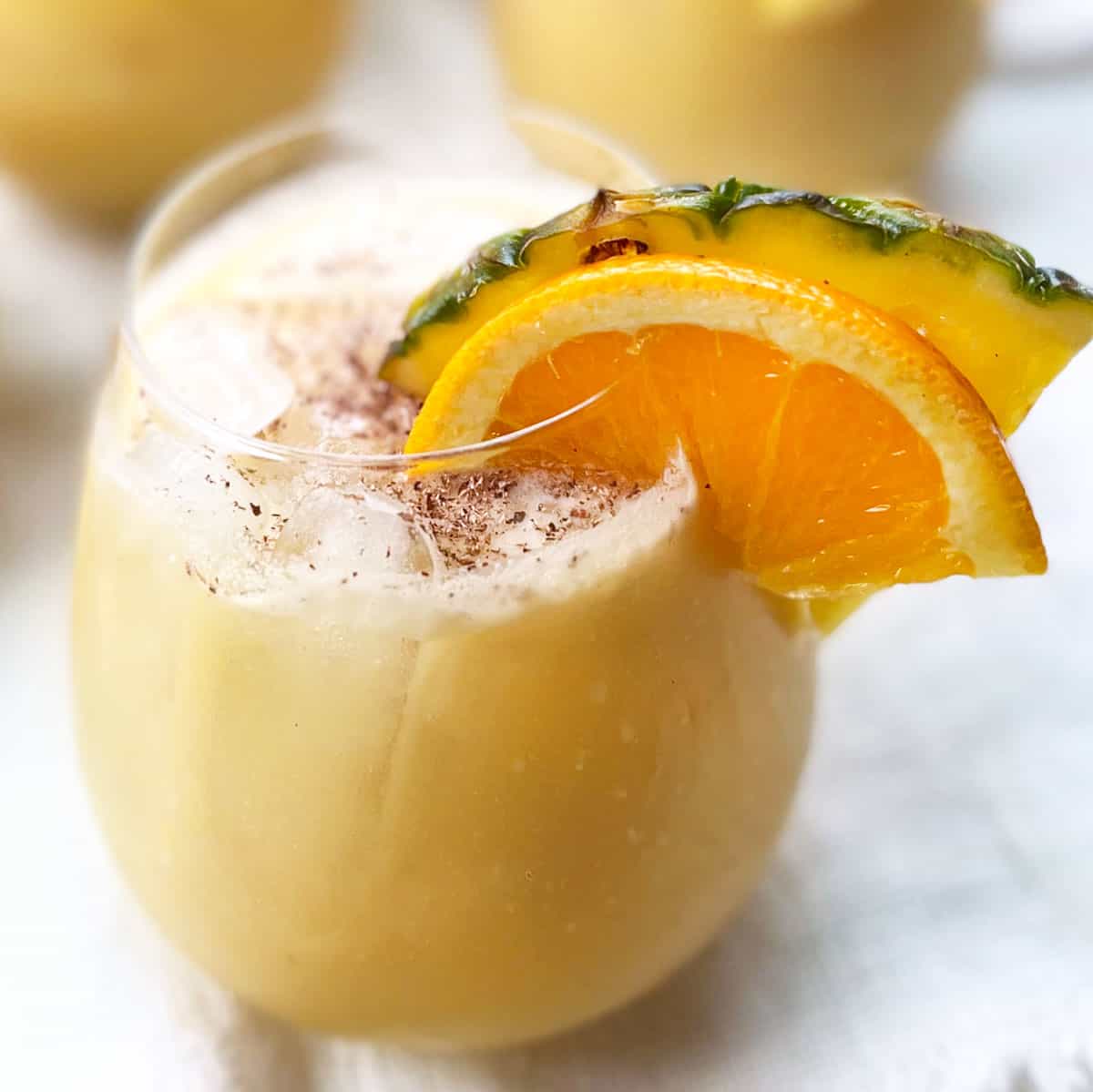 Close up of a creamy yellow painkiller cocktail on ice in a rounded cocktail glass with a wedge of orange and a wedge of pineapple attached to the rim