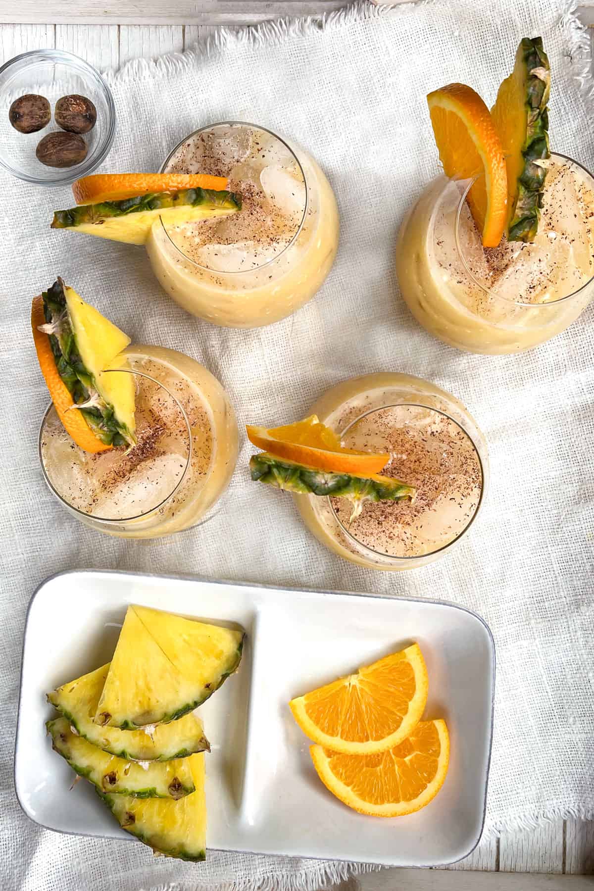 overhead shot of 4 large round cocktail glasses filled with painkiller cocktails on ice, each glass garnished with a slice of orange and a slice of pineapple, a white rectangular dish holds a few extra fruit slices.