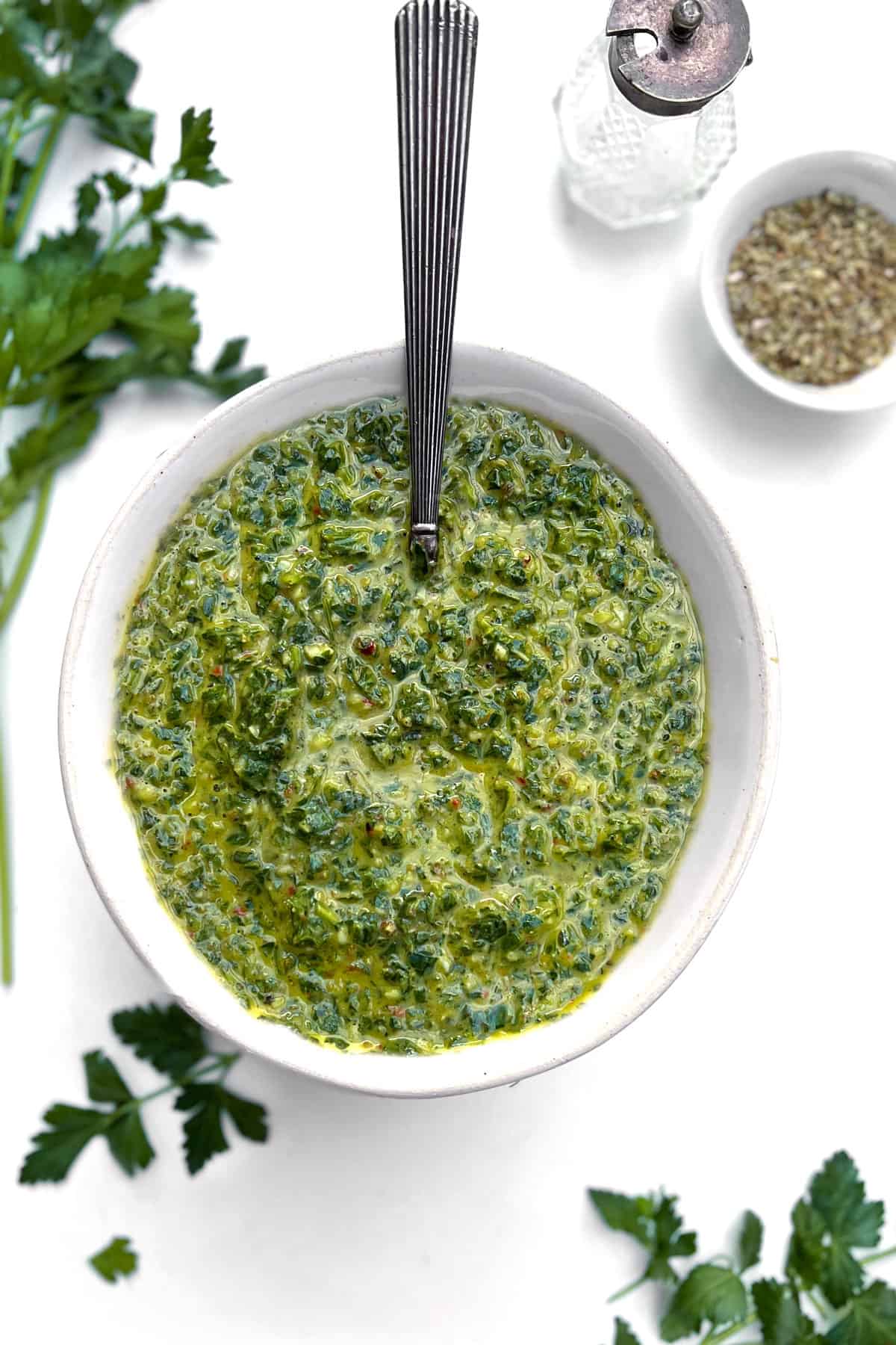overhead shot of a white bolw filled with green chimichurri sauce, a spoon lifting up some of the sauce, a small bowl of dried oregano and a salt shaker next to it.