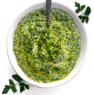 Overhead shot of a white bowl filled with vivid green chimichurri sauce, two parsley sprigs surrounding it