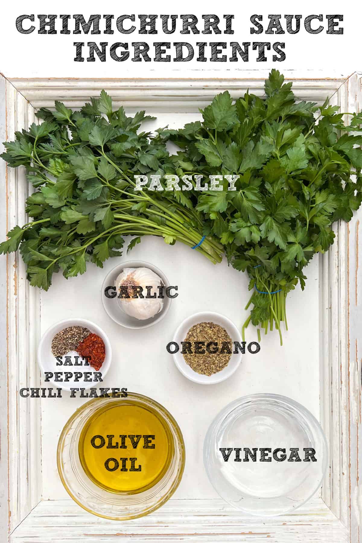 overhead shot of the 6 main ingredient in chimichurri sauce, a bunch of parsley, 5 garlic cloves, a small glass bowl of olive oil, a small glass bowl of white vinegar, tiny glass bowl of dried oregano and tiny glass bowl of salt, pepper and chili flakes
