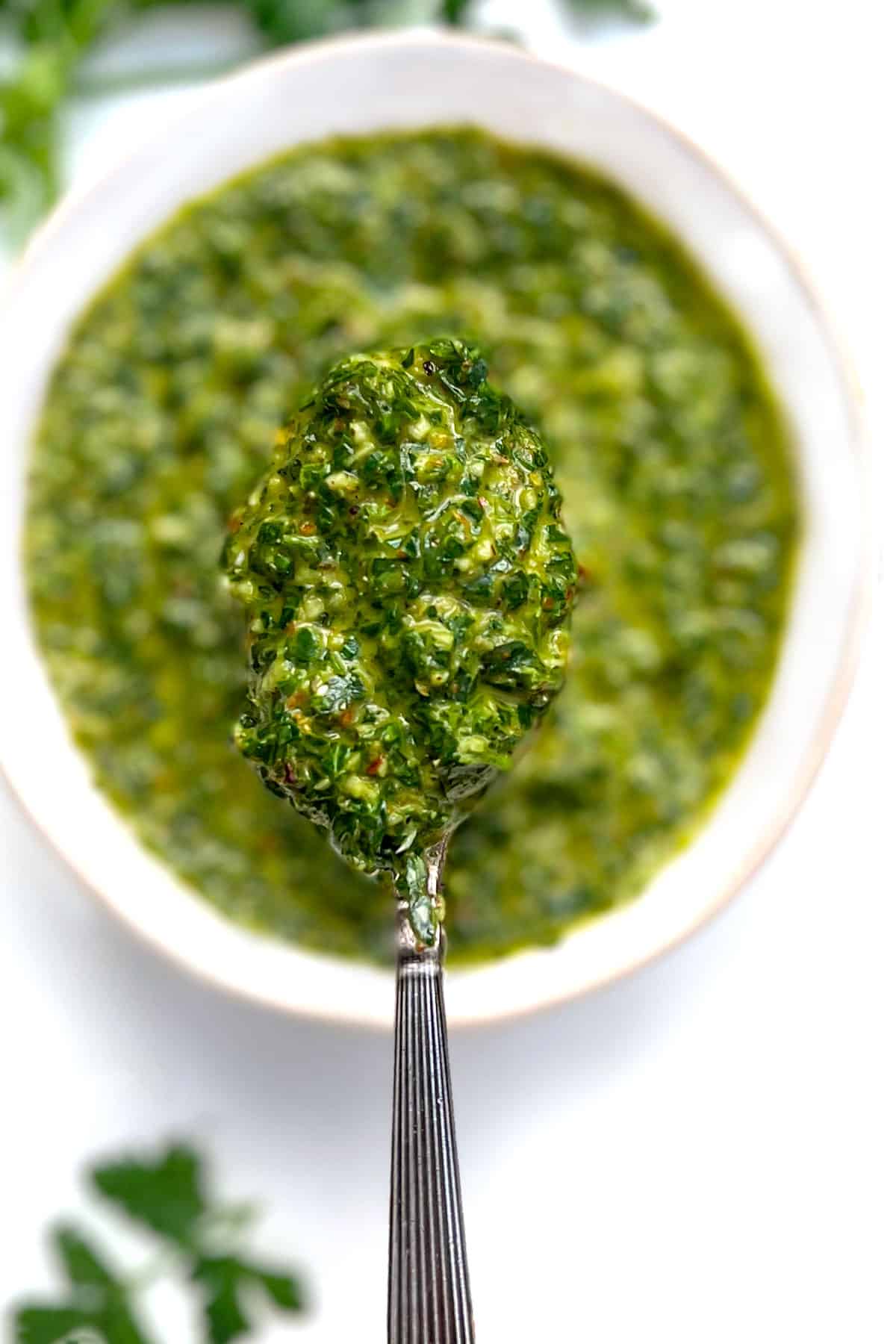 overhead shot of a white bolw filled with green chimichurri sauce, a spoon lifting up some of the sauce, a small bowl of dried oregano and a salt shaker next to it.