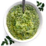 Overhead shot of a white bowl filled with vivid green chimichurri sauce, two parsley sprigs surrounding it