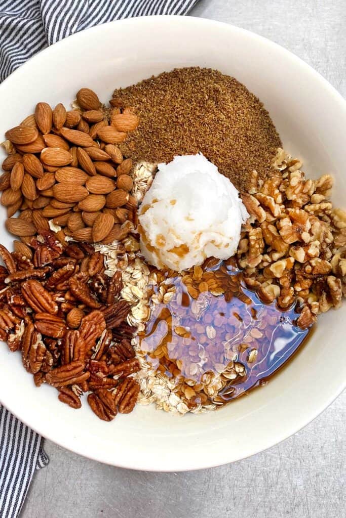 overhead shot of a white mixing bowl filled with granola ingredients in piles: oats, almonds, pecans, walnuts, ground flax, a blog of coconut oil in the middle and some maple syrup poured over the oats.