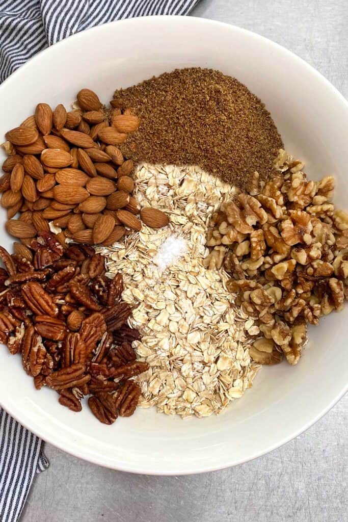 overhead shot of a white mixing bowl filled with granola ingredients in piles: oats, almonds, pecans, walnuts, ground flax and a pinch of salt.
