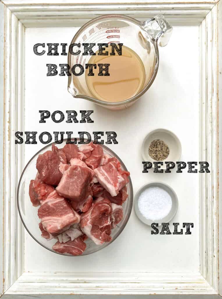 white wooden tray from above with glass bowl filled with cubes of raw pork shoulder and a smaller glass bowl filled with chicken broth and a tiny bowl of salt and a tiny bowl of pepper