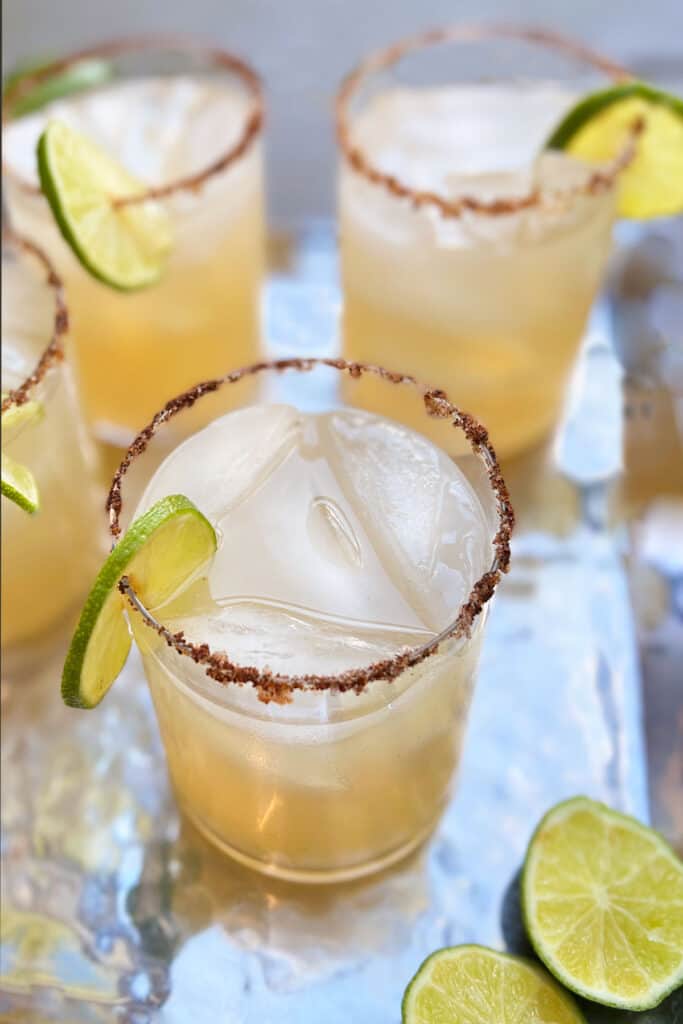 A cocktail glass filled with ice and a Mezcal cocktail mix of lime juice, simple syrup and sriracha. Glass is rimed with chili salt and has a wheel of lime on it.