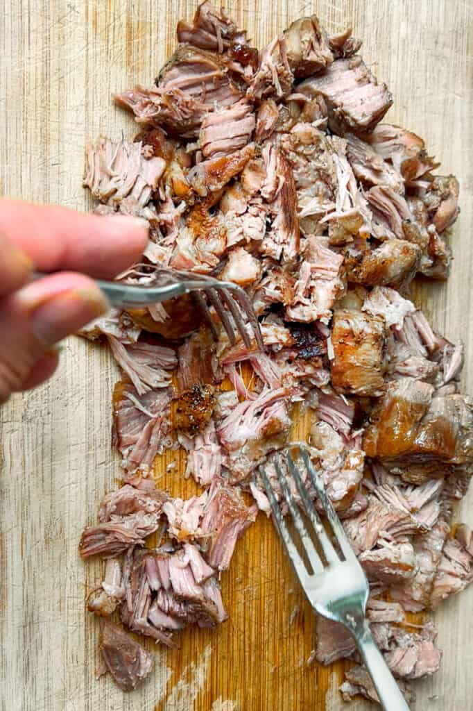 overhead shot of a wooden cutting board topped with pork carnitas that are being shredded with two forks