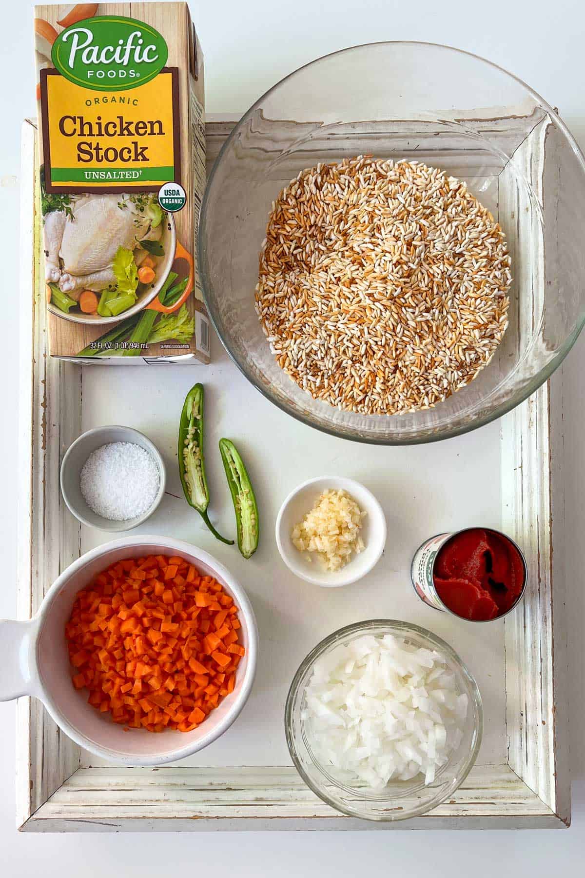 overhead shot of various ingredients for Mexican red rice (arroz rojo), on a white tray: small bowl of chopped white onion, small bowl of chopped carrots, tiny bowl of chopped garlic, open can of tomato paste, halved serrano chile pepper, large bowl of toasted jasmine rice and tiny bowl of salt