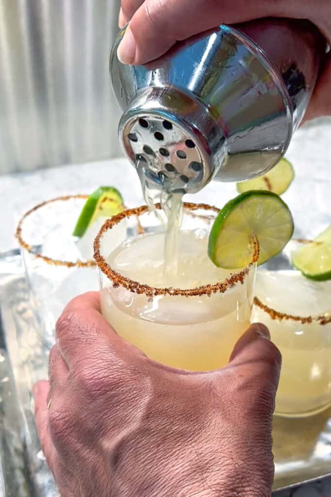 Hand holding a cocktail glass while another hand is pouring liquid into the glass through a cocktail shaker. Glass is rimmed with salt and chili powder and garnished with a slice of lime