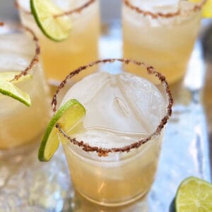 a cocktail glass filled with mezcal and lime juice and simple syrup and garnished with a ring of lime and a salt and chili rim, 3 other similar glasses in the background