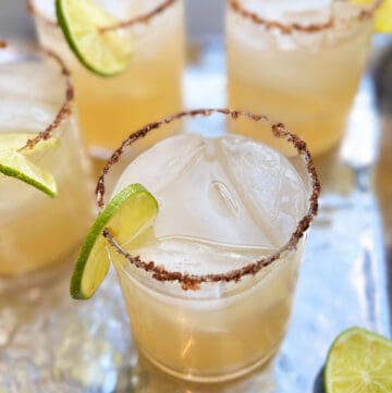 a cocktail glass filled with mezcal and lime juice and simple syrup and garnished with a ring of lime and a salt and chili rim, 3 other similar glasses in the background