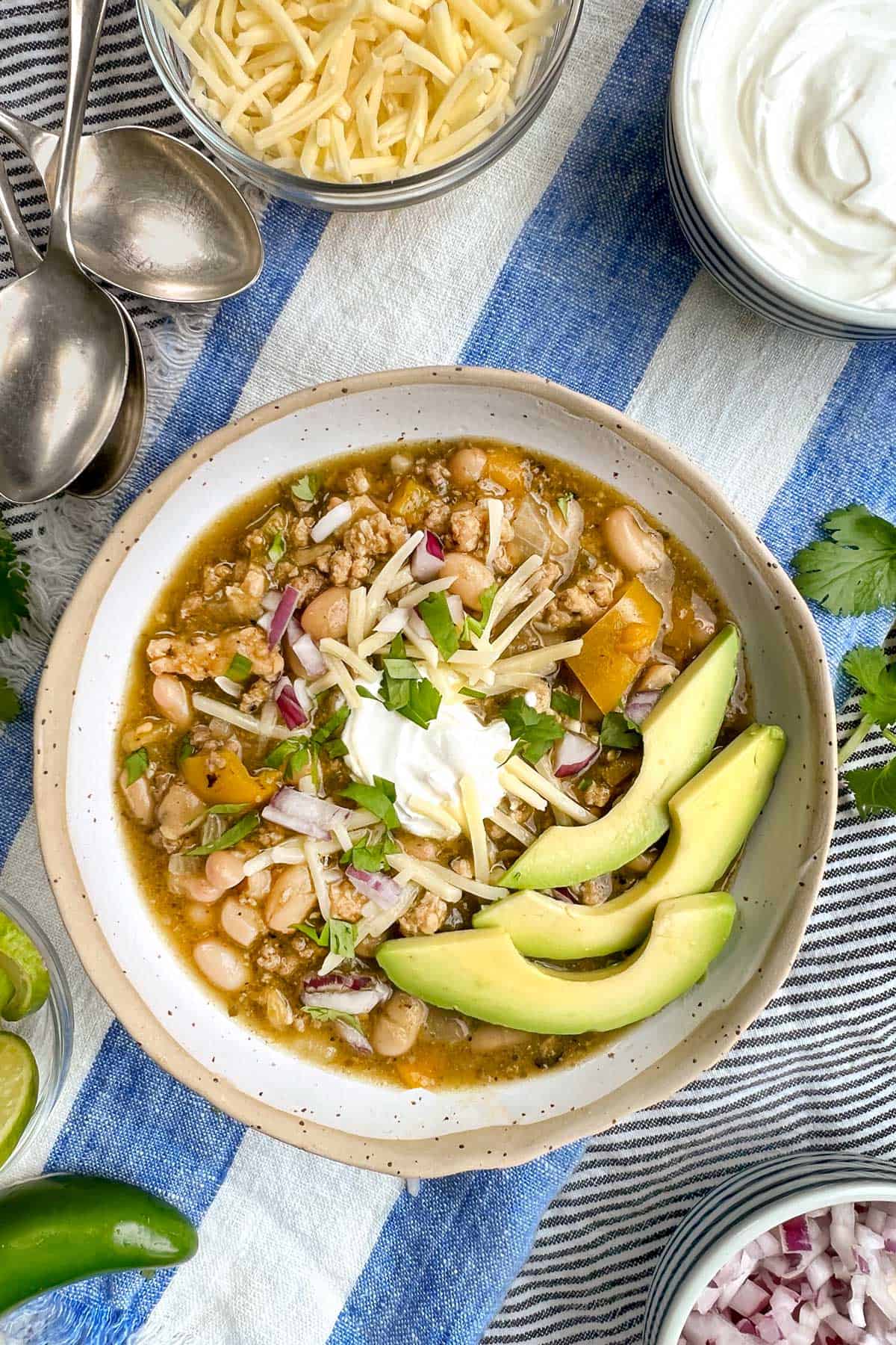 bowl of turkey chili garnished with sliced avocado, shredded cheese, sour cream and chopped cilantro, bowls of garnishes on the side