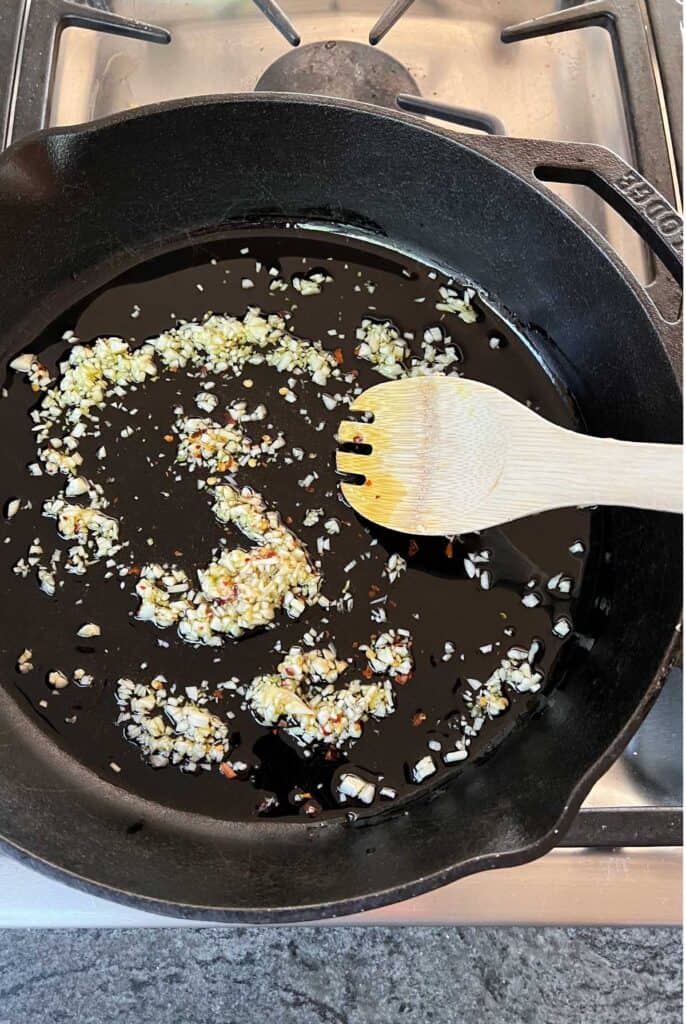 chopped garlic and red pepper flakes simmering in a cast iron skillet with a wooden spoon