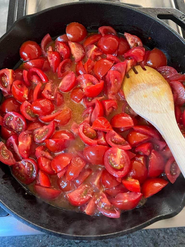 quartered sautéed cherry tomatoes with garlic and tomato juices, in a cast iron skillet with a wooden spoon.