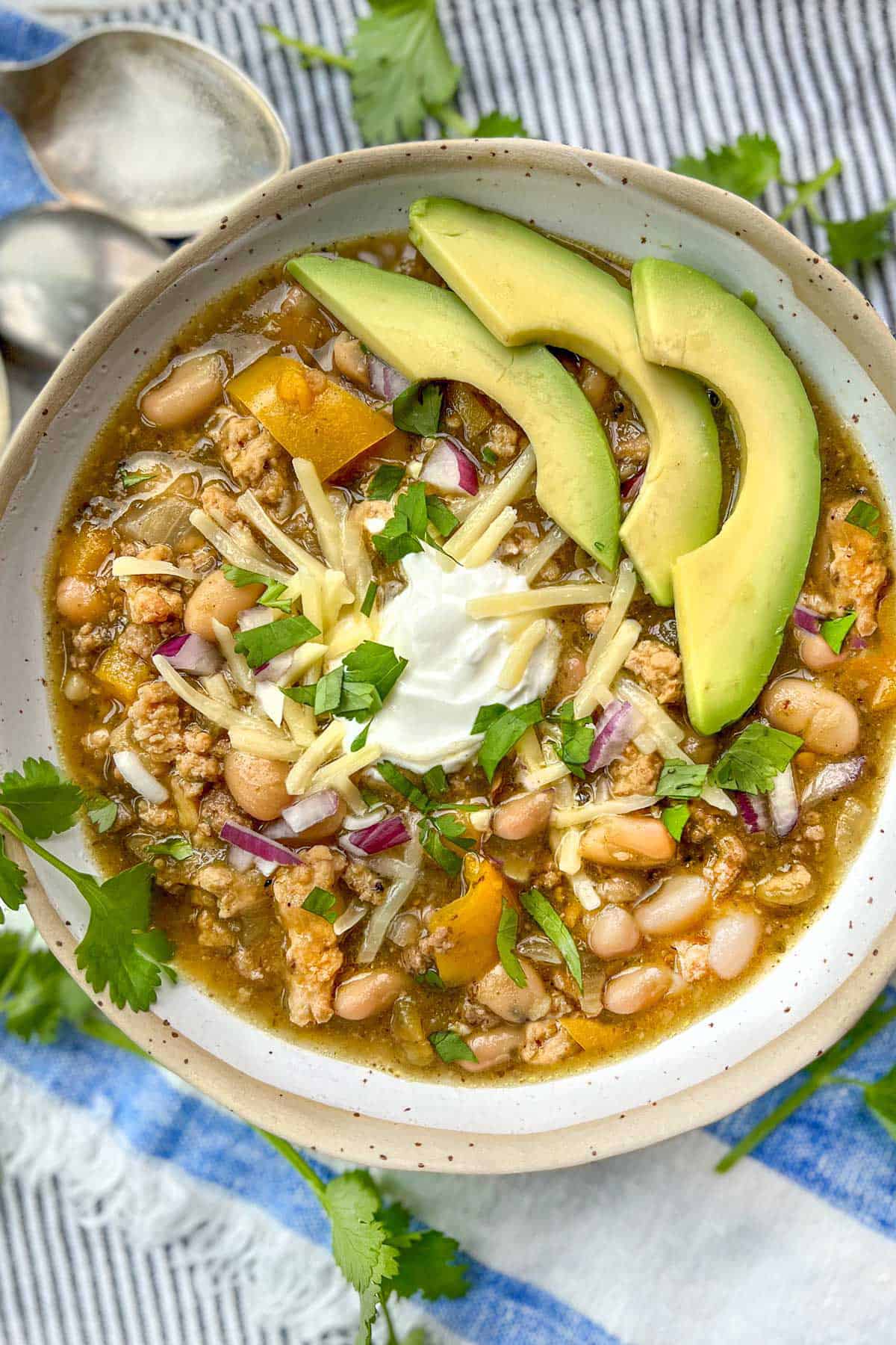 turkey white bean chili in a white bowl garnished with avocado, sour cream, red onions and shredded cheese