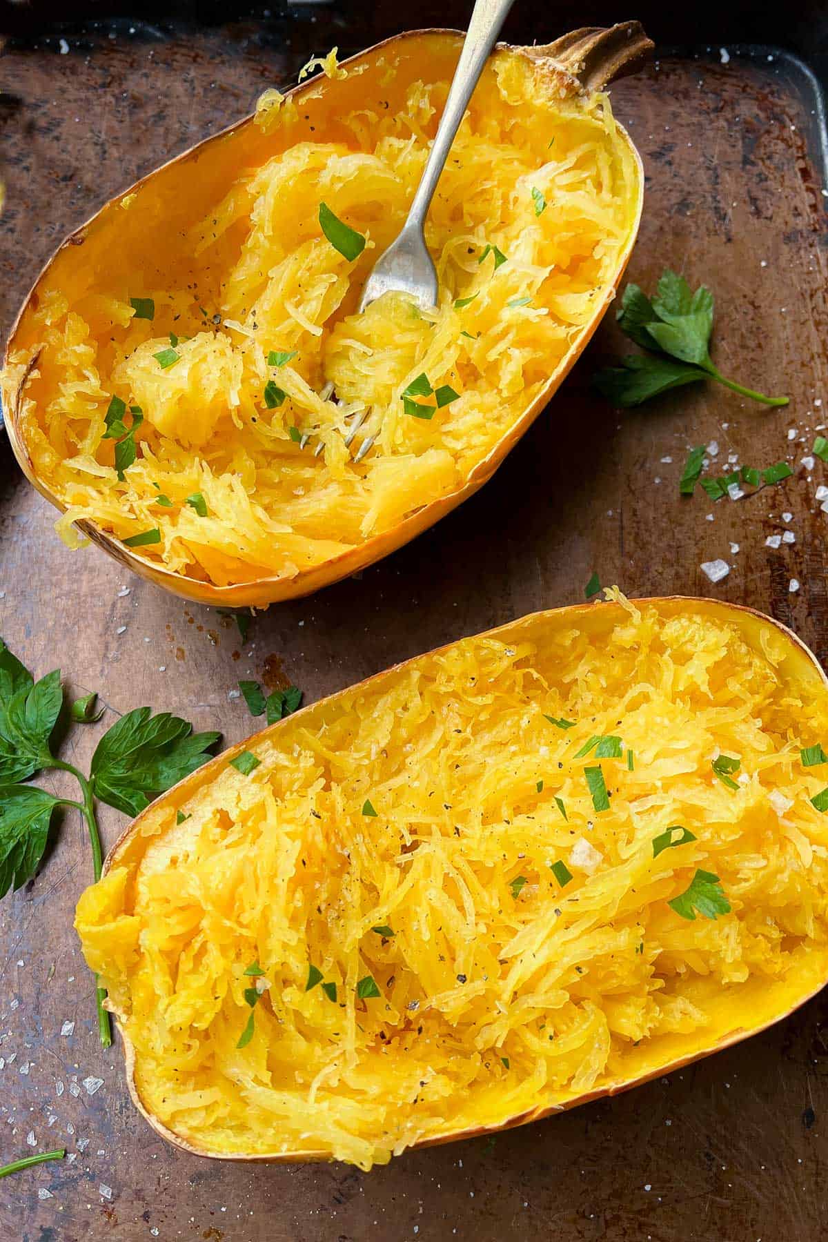 two roasted spaghetti squash halves with their strands pulled, fluffed and sprinkled with salt, pepper and parsley - a fork twirled into the strands of one squash half.