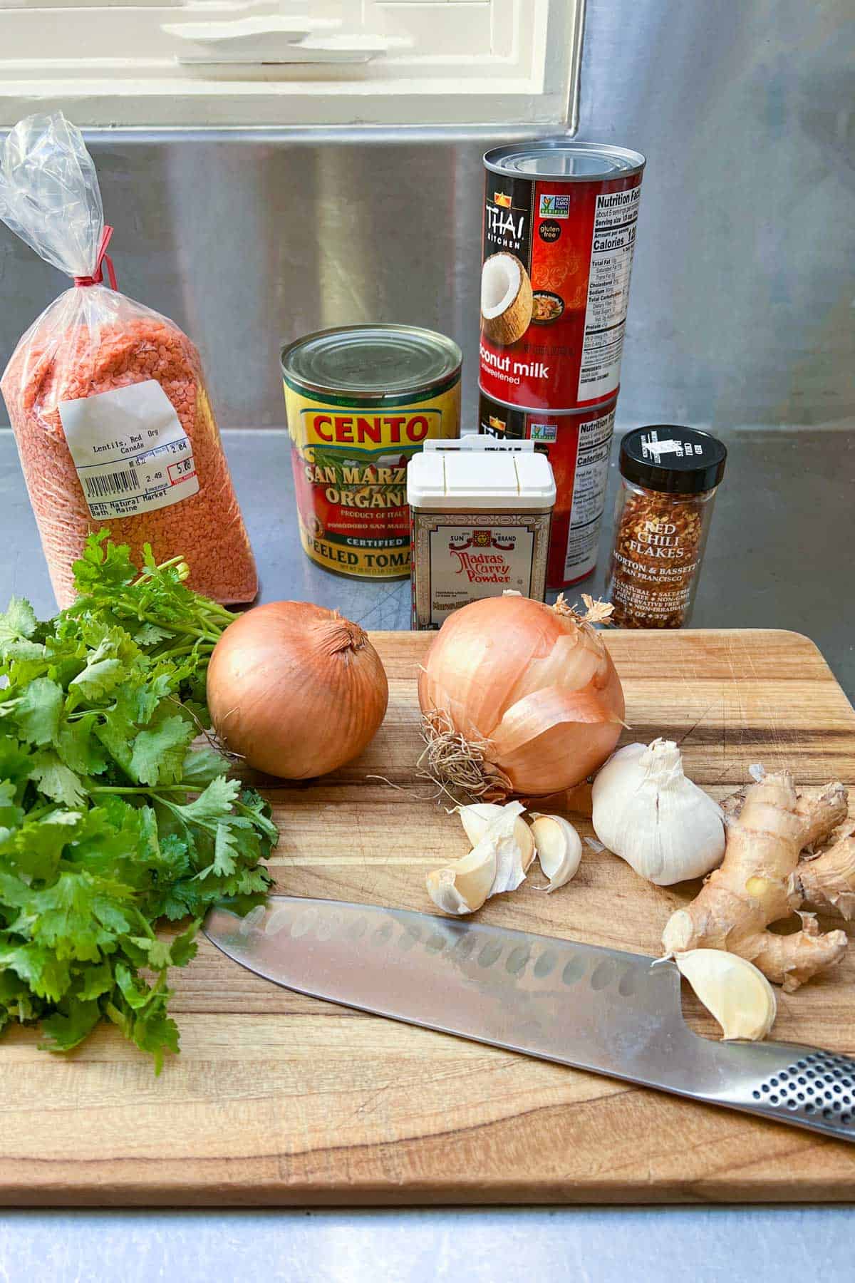 ingredients on a countertop: bag of red lentils, can of cento peeled tomatoes, container of madras curry powder, jar of red chili flakes, 2 cans of coconut milk, bunch of cilantro, 2 yellow onions, bulb of garlic, hunk of ginger root, kitchen knife.