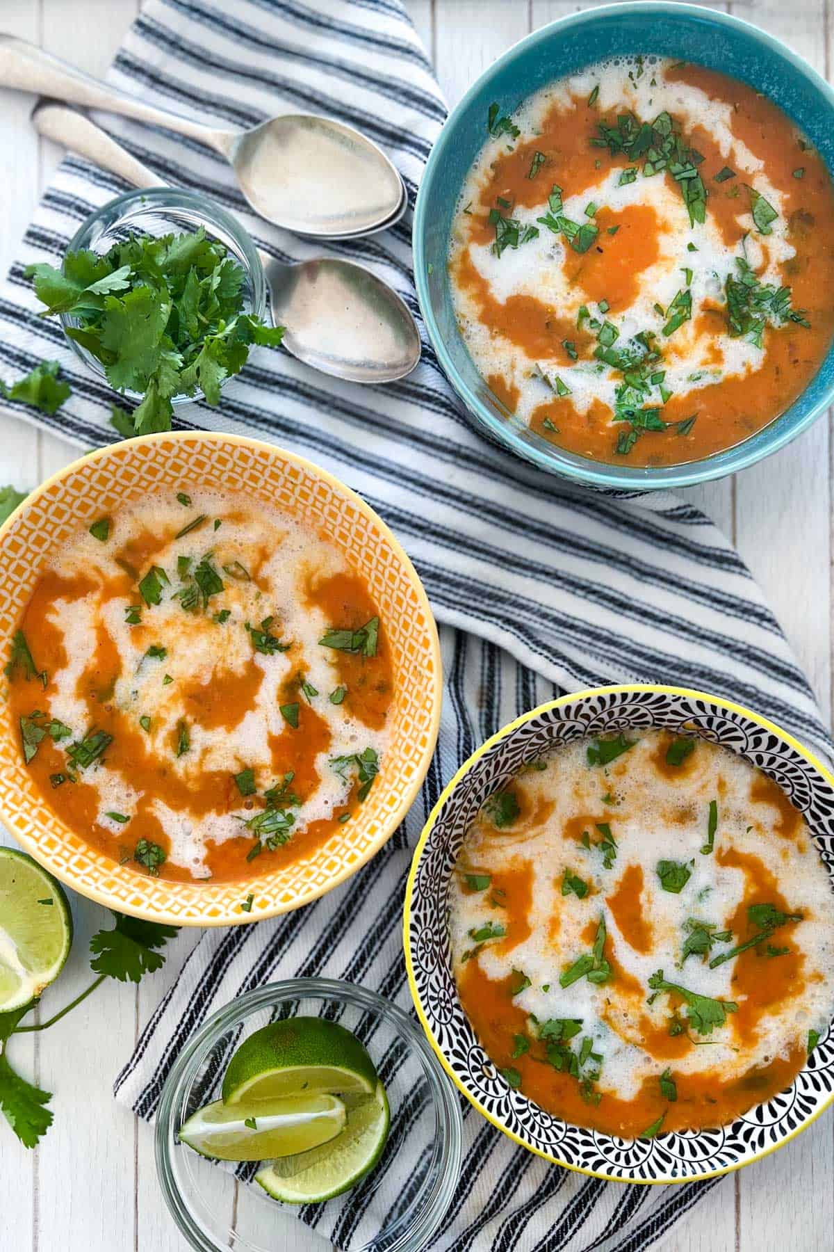 three bowls on a black and white striped dish towel, filled with red lentil soup drizzled with coconut milk and garnished with chopped cilantro