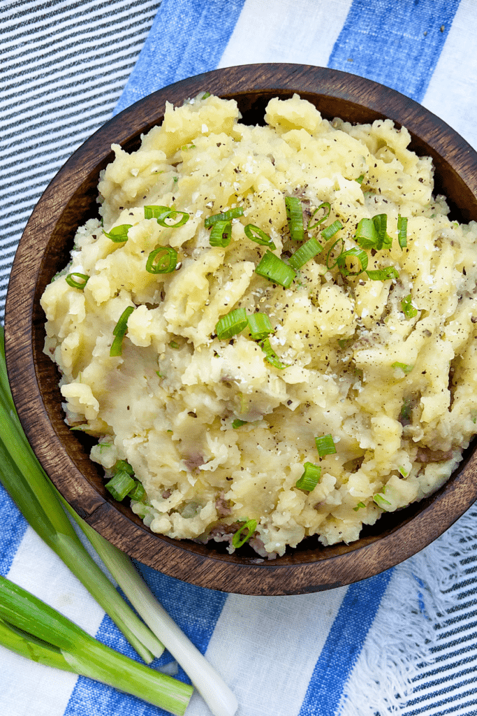 Wooden bowl filled with mashed red skin potatoes flecked with chopped scallions, on a blue and white striped cloth napkin