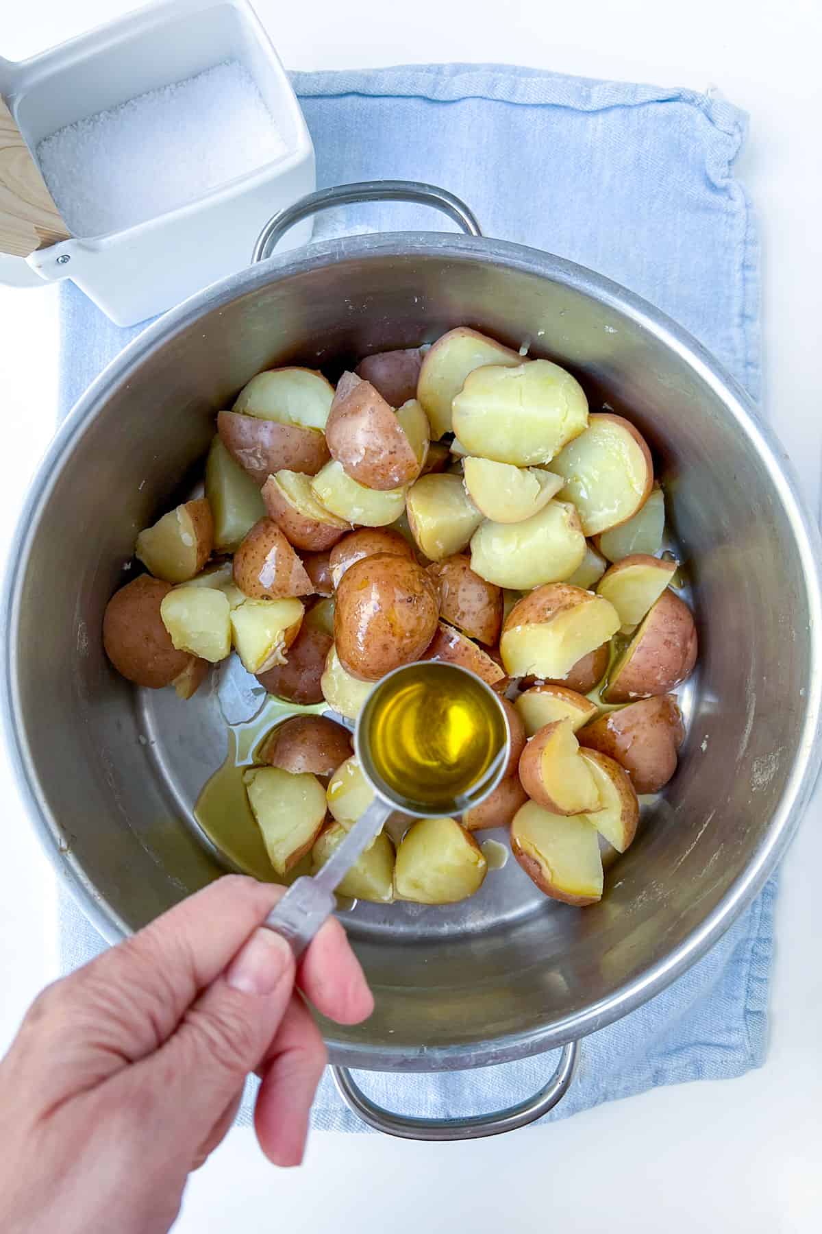 A hand holding a tablespoon of olive oil over a silver pot filled with boiled quartered red skin potatoes.