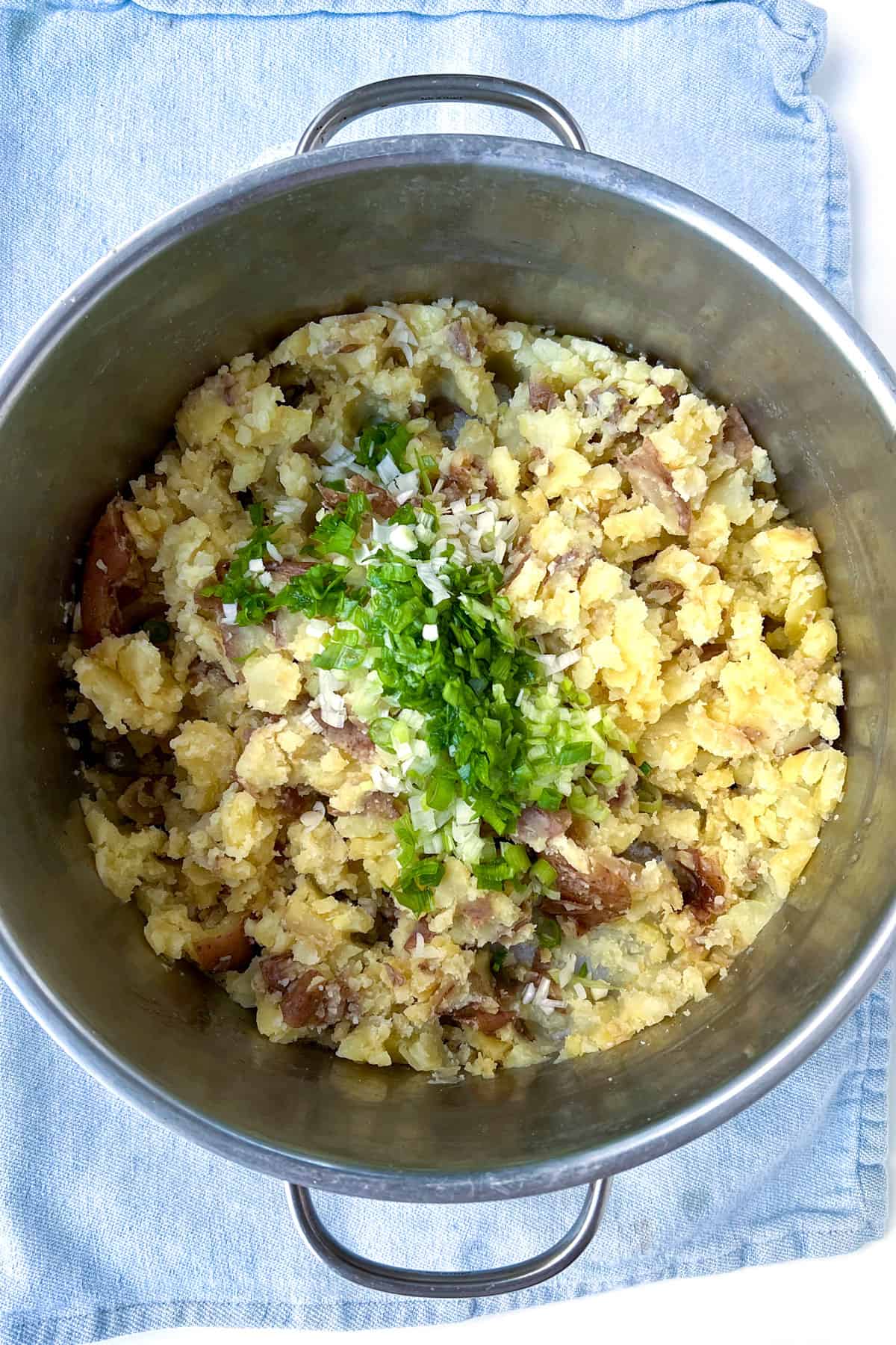 Chopped scallions on top of roughly mashed red skin potatoes, in a silver cooking pot, on a blue kitchen towel