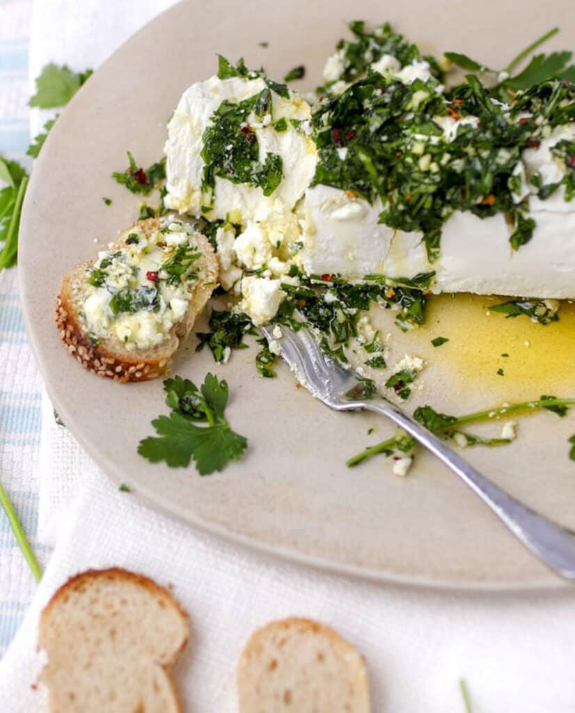 a log of goat cheese topped with parsley garlic dressing on a tan plate, with one baguette slice topped with the goat cheese and a small serving knife nearby.