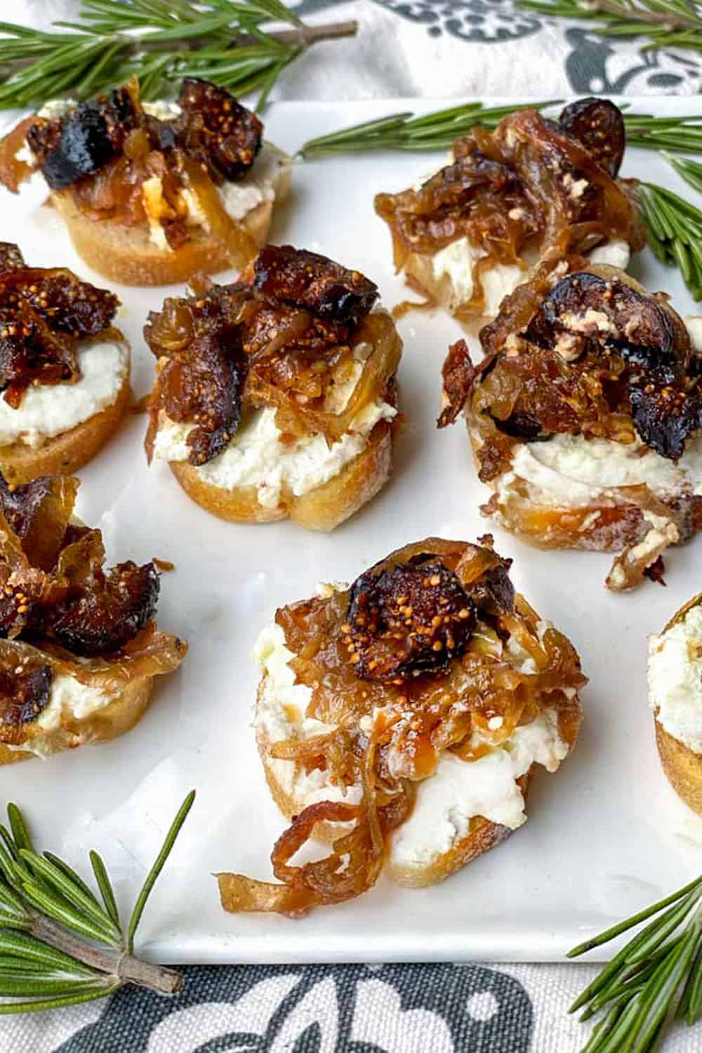 7 baguette slices on a white serving plate topped with goat cheese and caramelized onions mixed with figs, rosemary sprigs for garnish