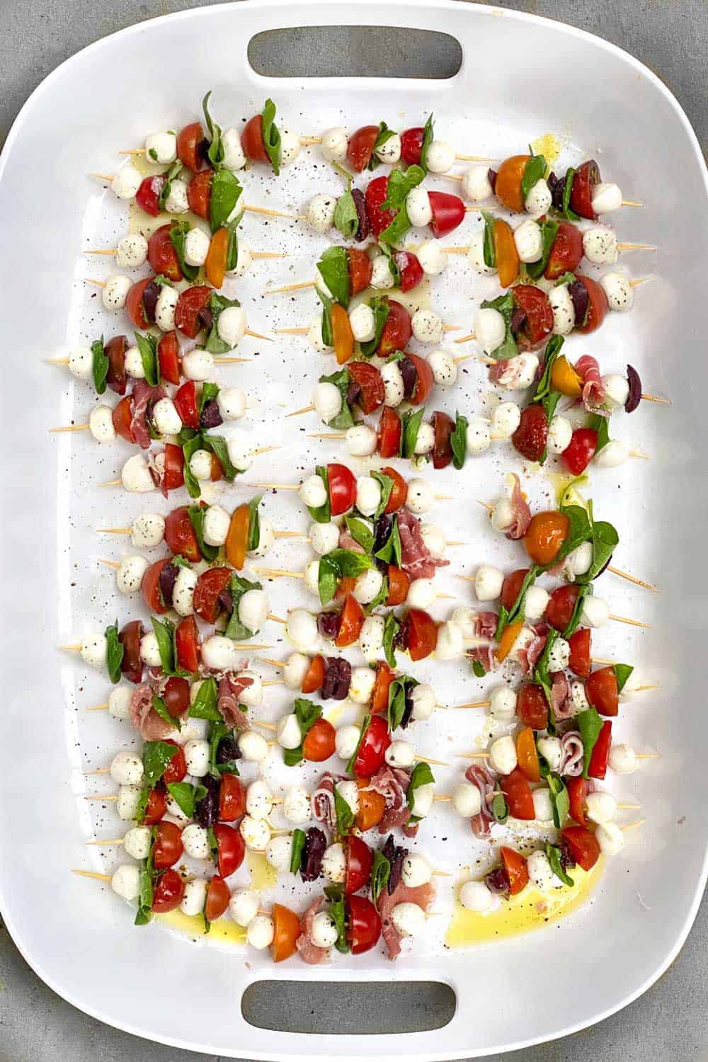 White serving tray filled with dozens of mini caprese kabobs, each one with 3 mini mozzarella balls, two halved cherry tomatoes and a couple of folded basil leaves, some with added prosciutto and sliced kalamata olives.