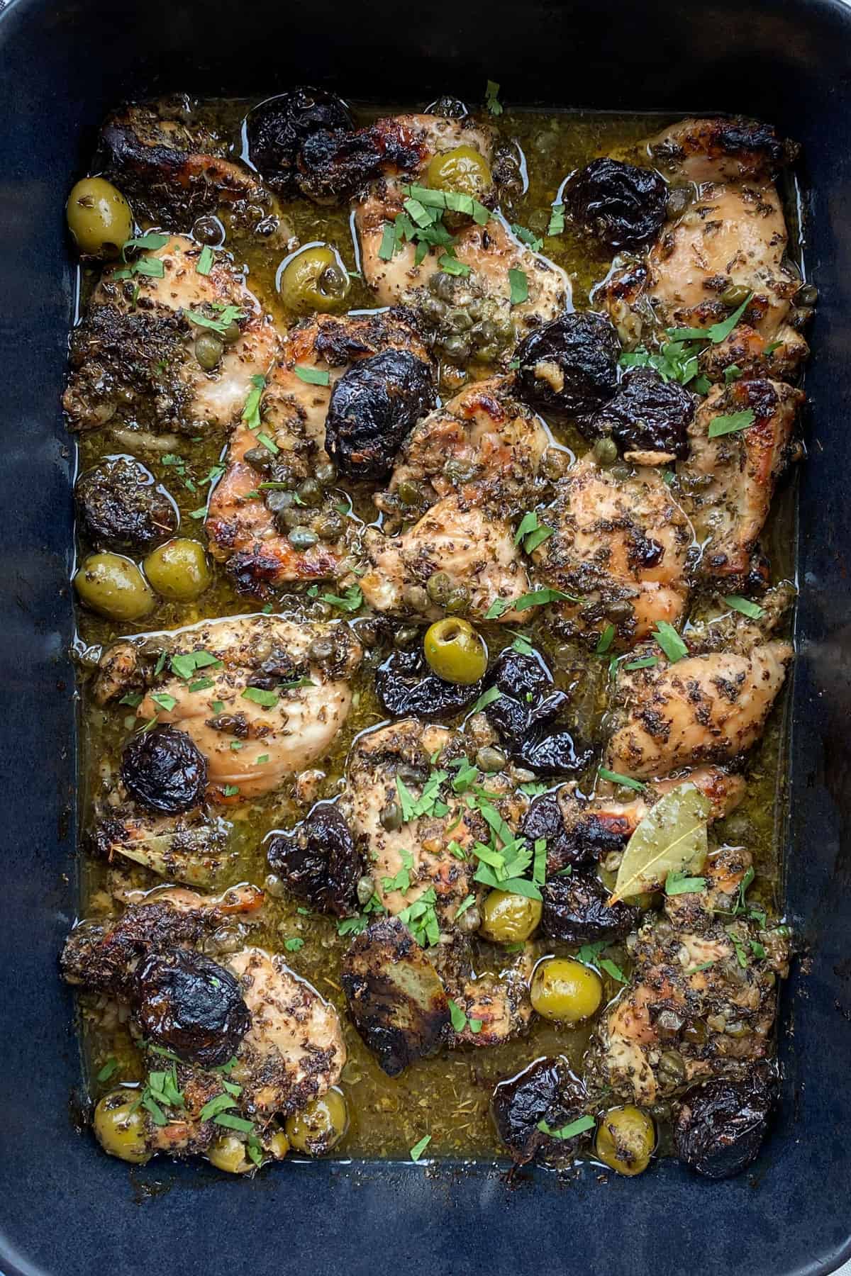 10 baked boneless chicken thighs in a black rectangular casserole pan, soaking in pan juices with prunes and olives and a garnish of chopped parsley.