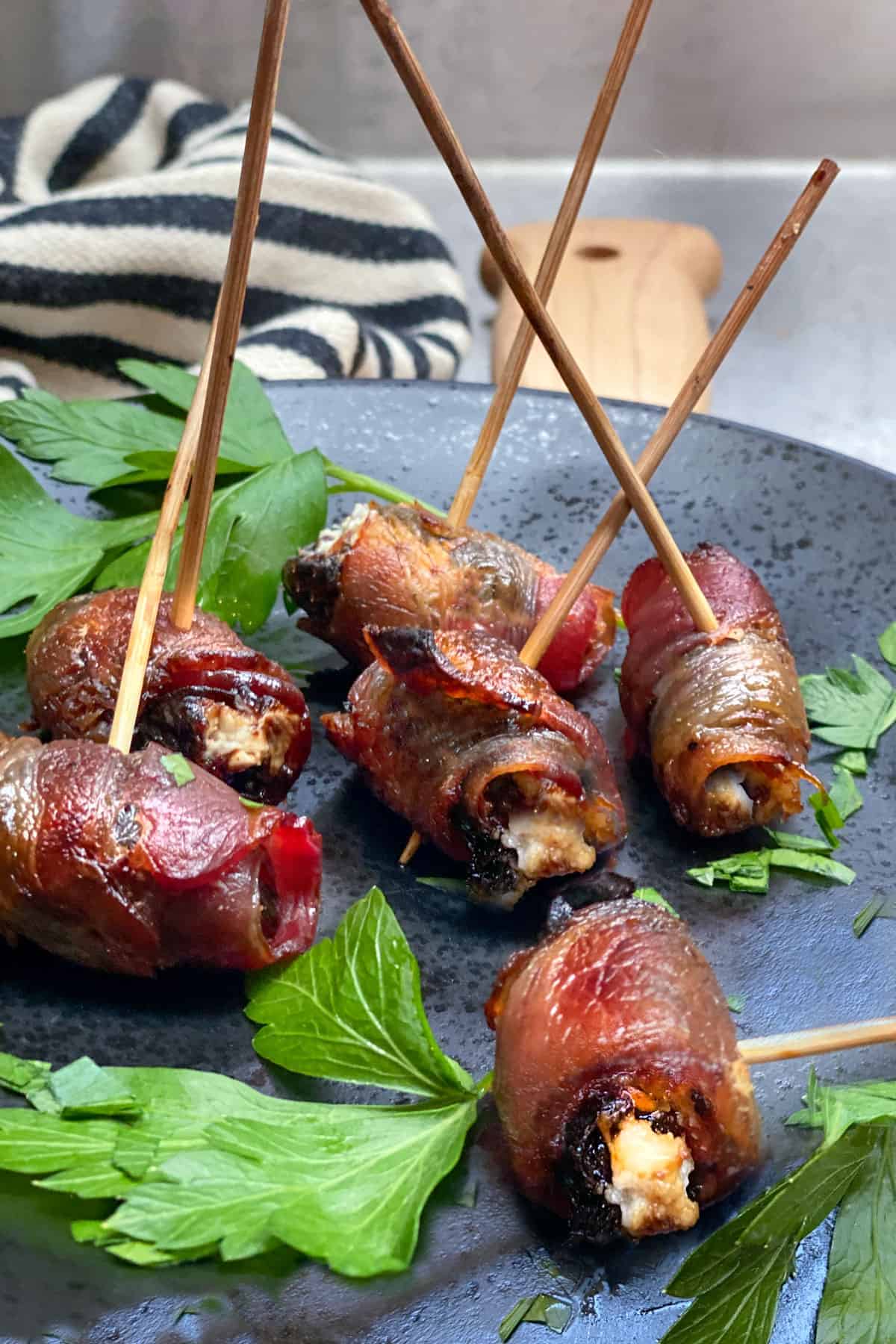 6 bacon wrapped, goat cheese stuffed prunes on a black plate with long bamboo skewers stabbed into each one
