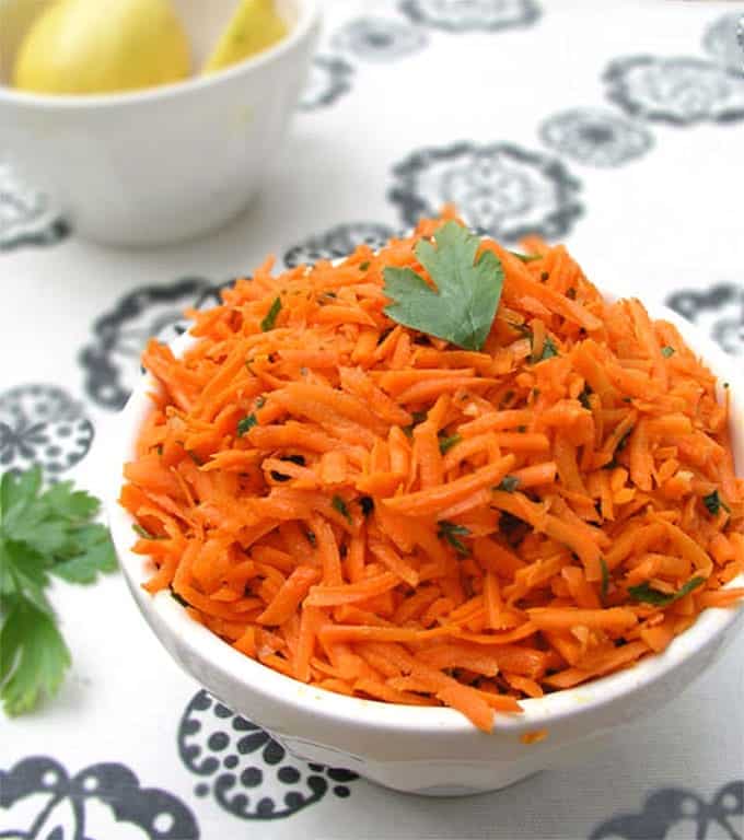 white bowl sitting on a decorative black and white tablecloth, filled with shredded carrot salad that's flecked with chopped parsley.