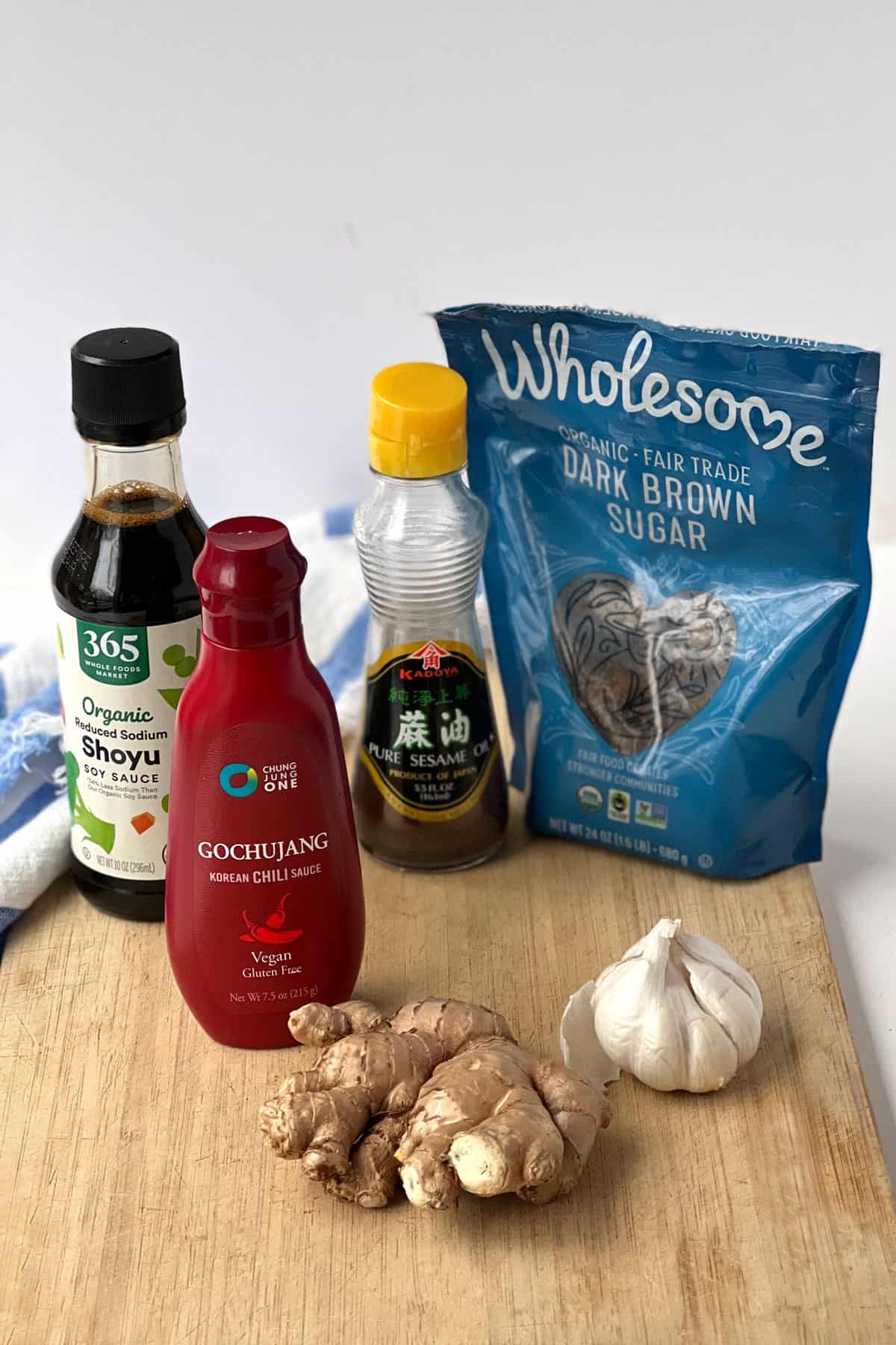 ingredients for gochujang sauce on a wooden cutting board: bottle of soy sauce, bottle of gochujang sauce, bottle of sesame oil, bag of dark brown sugar, whole bulb of garlic, hunk of ginger root