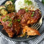 two slow cooked chicken thighs with vivid orange gochujang sauce on a black plate with some white rice and a few slices of sauteed zucchini.