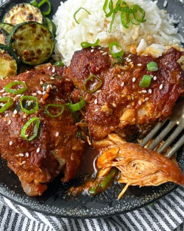two slow cooked chicken thighs with vivid orange gochujang sauce on a black plate with some white rice and a few slices of sauteed zucchini.