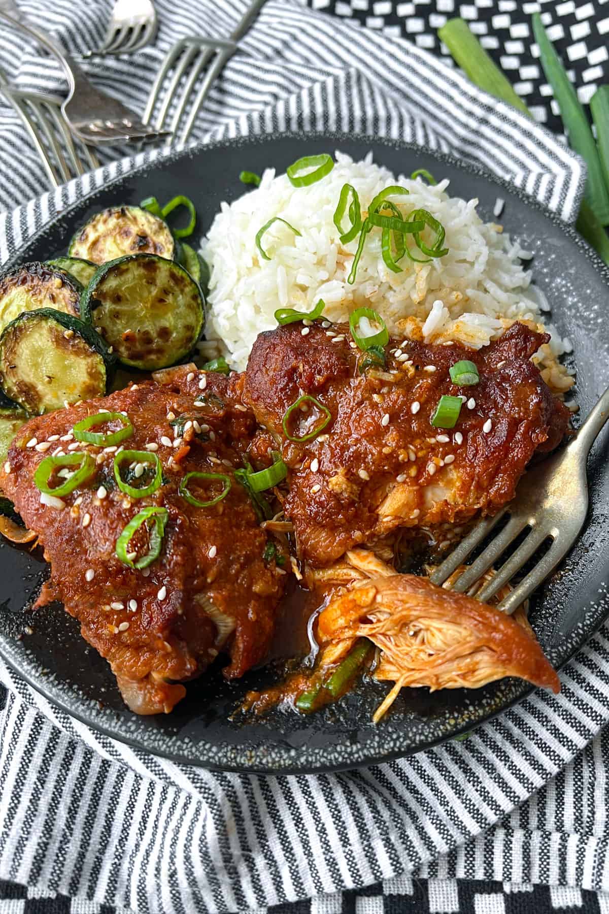 two slow cooked chicken thighs with vivid orange gochujang sauce on a black plate with some white rice and a few slices of sauteed zucchini.