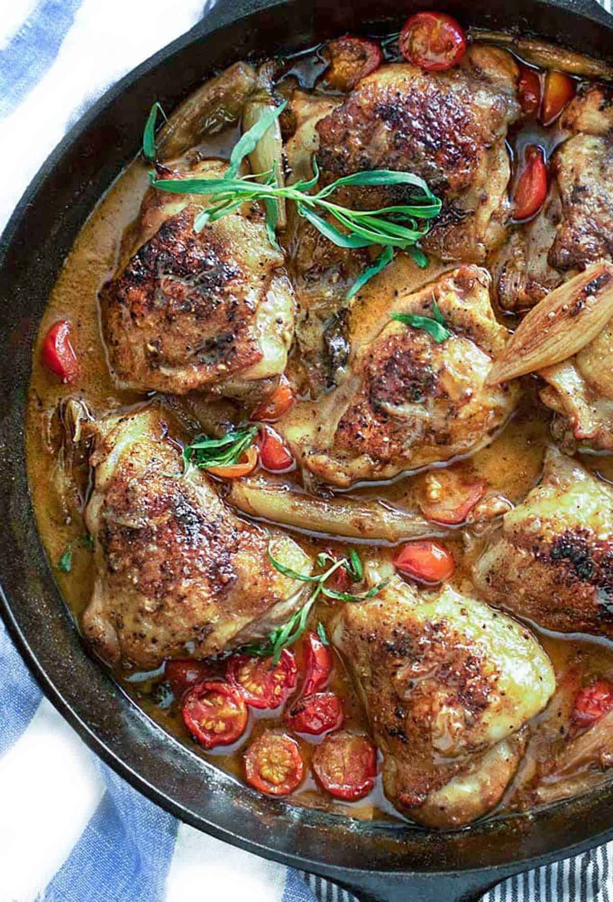 cast iron skillet filled with 8 braised chicken thighs in a brown sauce with cherry tomatoes and shallots