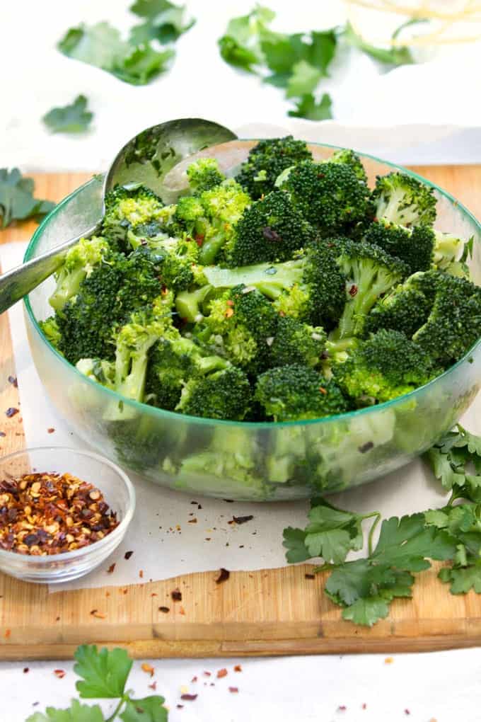 broccoli florets in a clear green glass bowl with a small bowl of crushed red pepper flakes in the foreground and some parsley leaves strewn around.
